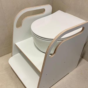 Pode incluir: Um degrau de treino sanit&aacute;rio branco com design de dois n&iacute;veis e pegas laterais, constru&iacute;do em madeira com acabamento branco e bordas de contraplacado expostas. O texto WHITE MEL est&aacute; impresso a preto acima da unidade que rodeia o assento sanit&aacute;rio.