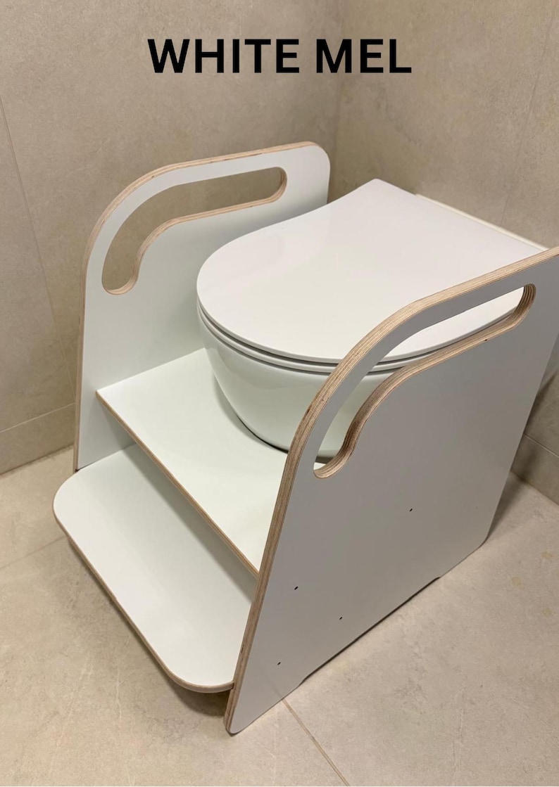 Pode incluir: Um degrau de treino sanit&aacute;rio branco com design de dois n&iacute;veis e pegas laterais, constru&iacute;do em madeira com acabamento branco e bordas de contraplacado expostas. O texto WHITE MEL est&aacute; impresso a preto acima da unidade que rodeia o assento sanit&aacute;rio.