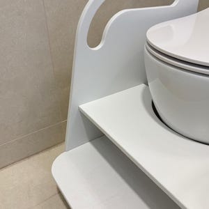 Pode incluir: Um banco de degrau de madeira branco com acabamento liso e uma al&ccedil;a recortada no topo. A estrutura apresenta uma plataforma plana e um painel lateral curvo. A palavra WHITE est&aacute; impressa em preto no topo da estrutura, perto da al&ccedil;a.