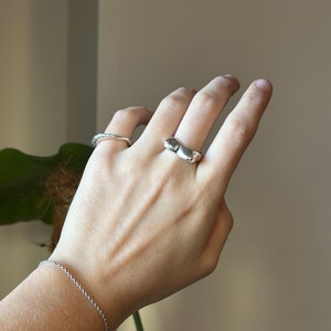May include: A silver ring with a unique, organic shape. The ring is worn on the middle finger of a hand, along with a thin silver band on the ring finger.