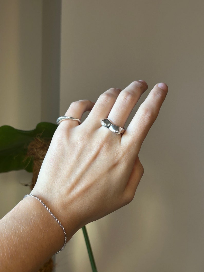 May include: A silver ring with a unique, organic shape. The ring is worn on the middle finger of a hand, along with a thin silver band on the ring finger.