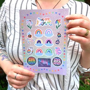 Könnte beinhalten: Ein Bogen mit bunten Aufklebern mit Regenbogen, Blumen, Vögeln, Bienen und Marienkäfern. Die Aufkleber tragen die Aufschriften "we are love", "safe space" und "protect the dolls". Die Aufkleber befinden sich auf einem lavendelfarbenen Hintergrund mit Glitzer.