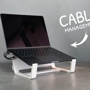 May include: A white laptop stand holding a black laptop with a dark screen. The stand features a built-in cable management system. White text on the right side reads CABLE MANAGEMENT with a white arrow pointing toward the stand base.