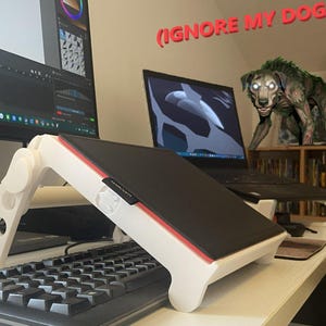 Peut inclure: Un support pour ordinateur portable blanc réglable avec une surface noire et une bordure rouge, doté d'un design à charnière mécanique avec des découpes circulaires. Le support est placé au-dessus d'un clavier d'ordinateur noir.