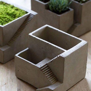May include: A set of gray concrete planters featuring a multi-level design with integrated miniature staircases. One planter is empty, while the others contain green succulents and moss, showcasing a geometric structure with distinct compartments.