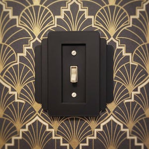 May include: A matte black light switch plate with a stepped, tiered rectangular design. The plate features a central vertical toggle switch in a light cream color, secured by two flat-head screws positioned above and below the toggle mechanism.