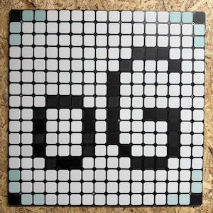 以下が含まれることがあります： 白い背景に黒い文字「ds」のピクセル化されたデザインが施された正方形のグリッドディスプレイボード。ボードは小さな丸みを帯びた正方形のタイルで構成されており、四隅には水色のアクセントタイルが配置されています。