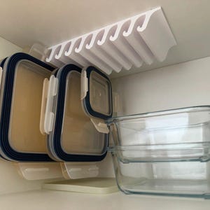 Puede incluir: Un organizador de cocina de plástico blanco montado bajo un estante de armario, que sostiene varios recipientes rectangulares con sellos de silicona azul oscuro y tapas transparentes, junto a recipientes de vidrio apilados.