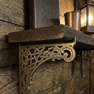 May include: A dark brown wooden shelf supported by an ornate brass-coloured metal bracket featuring intricate Celtic knotwork and a dragon motif. The shelf holds a dark brown book with a Celtic design on the cover and a hammered copper-coloured cup.