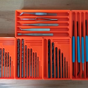 May include: A collection of metal tools organized in bright orange plastic trays. The set includes various silver tweezers, multiple black drill bits of graduated sizes, and several long tools with blue handles arranged in vertical slots.