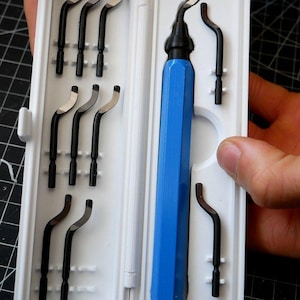 May include: A blue deburring tool with a hexagonal handle and a set of eight interchangeable black metal blades, all organized within a white plastic storage case. The tool features a curved blade attached to the handle, while seven additional blades are secured in molded slots inside the case.