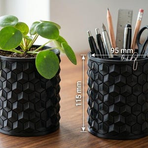 May include: Two black cylindrical containers with a textured geometric hexagonal pattern. One holds a green Pilea plant, while the other contains office supplies like pens, a ruler, and scissors. Dimensions are 95 mm in diameter and 115 mm in height.