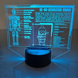 May include: A blue-glowing acrylic IG-88 Assassin Droid lamp. The lamp features a detailed diagram of the droid and informational text. The base is a black cylinder. The text includes "IG-88 ASSASSIN DROID".