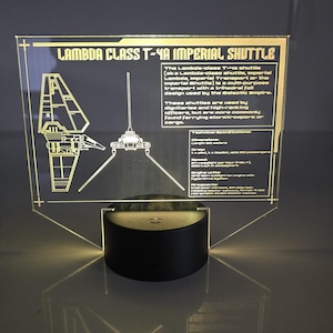 May include: A Lambda Class T-4a Imperial Shuttle illuminated display. The design is etched in gold on a clear acrylic panel, with a black base. The text on the panel describes the shuttle's features and use by the Galactic Empire.