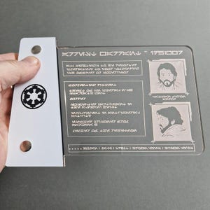 May include: A black and white ID card with a white background and black text. The text includes the words "K77K" and "1751007". The card has two holes at the top and a small image of a symbol at the bottom left corner.