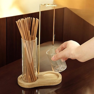 Puede incluir: Un soporte de madera con dos recipientes de vidrio. Un recipiente contiene un paquete de palillos de madera, y el otro recipiente está vacío. Un soporte de metal dorado con un gancho sostiene un solo palo de incienso.