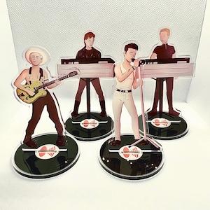 Puede incluir: Soportes acrílicos con una banda de cuatro músicos. Uno toca una guitarra amarilla, otro canta en un micrófono y dos tocan teclados. Cada figura se encuentra sobre una base negra con un patrón de camuflaje.