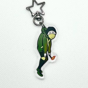 以下が含まれることがあります： 星型の金属製留め具が付いた緑と黒の漫画キャラクターのキーホルダー。キャラクターは緑色のジャケット、黒いズボン、黒いマスクを着用しています。キャラクターは火のついたマッチを持っています。