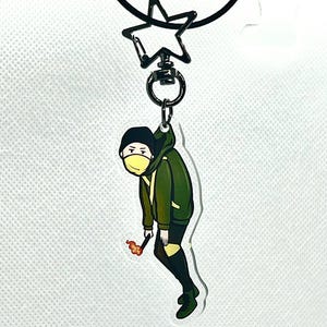 以下が含まれることがあります： 黄色のマスクと火のついたマッチを持った緑と黒の漫画のキャラクターのキーホルダー。キーホルダーは黒い星型のリングに取り付けられています。