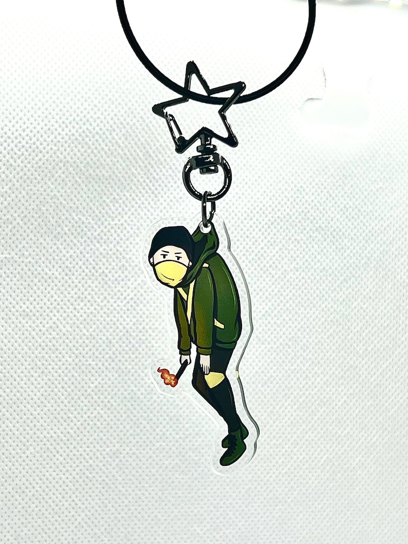 以下が含まれることがあります： 黄色のマスクと火のついたマッチを持った緑と黒の漫画のキャラクターのキーホルダー。キーホルダーは黒い星型のリングに取り付けられています。