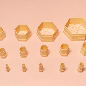 May include: A selection of light beige, hexagonal containers of different sizes, set against a pale pink backdrop. Some containers feature an internal lattice design. The containers are stacked and nested, implying a storage or organisational theme.