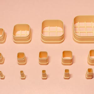 May include: A selection of beige square cookie cutters of varying sizes, displayed on a pale pink surface. Some cutters feature a grid design. The set includes different heights and widths, ideal for baking.