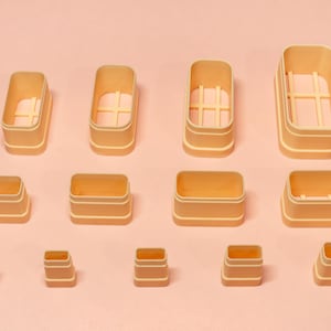 May include: An assortment of beige rectangular containers of different sizes, displayed on a pale pink surface. Some containers feature internal cross-shaped supports. The containers are empty and appear to be made of plastic or a similar material.