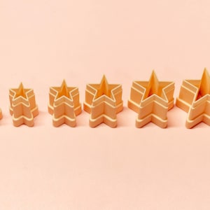 May include: A collection of six star-shaped cookie cutters in different sizes. The cutters are a light peach colour with white trim, set against a matching peach background. The star shapes have a three-dimensional design.