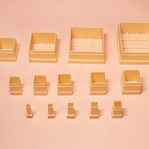 May include: A selection of beige square and rectangular containers of different sizes, arranged on a pale pink surface. Some containers feature internal grid structures. The containers are made of a solid material and are evenly spaced.