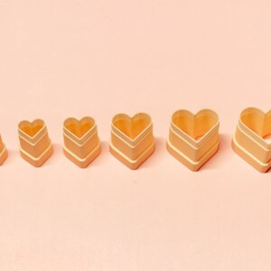 May include: A set of six heart-shaped cookie cutters in different sizes, displayed on a pink surface. The cutters are light orange with white detailing. These baking tools are perfect for creating heart-shaped treats, with sizes increasing from left to right.