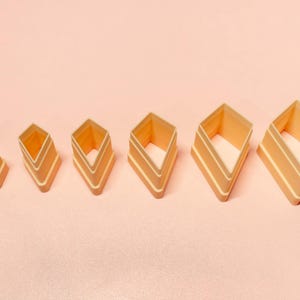 May include: A set of six diamond-shaped cookie cutters in varying sizes, arranged from smallest to largest. The cutters are a light orange color with white accent lines, set against a soft pink background.