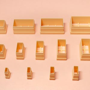 May include: A collection of beige rectangular containers of varying sizes, arranged on a light pink surface. The containers are open-topped, with some featuring internal cross supports. The overall aesthetic is minimalist and geometric.