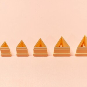 May include: Six peach-coloured, triangular-shaped cookie cutters of varying sizes are arranged in a row. Each cutter has a geometric design with a flat base and a pointed top. The cutters are on a light peach background.