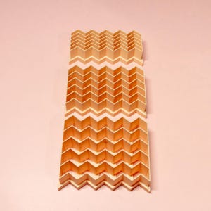 May include: A folded paper sculpture with a repeating zigzag pattern. The paper is a warm, light brown color with white accents. The sculpture is displayed against a soft pink background, creating a minimalist aesthetic.
