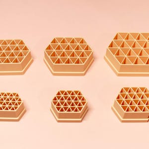 May include: Six peach-coloured, hexagonal organisers with a grid-like interior, arranged on a light pink background. The organisers vary in size, showcasing a range of storage options for small items.