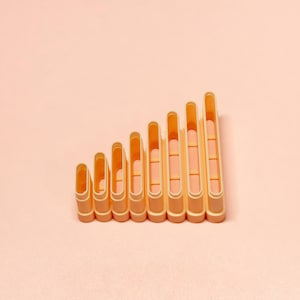 May include: Eight peach-coloured plastic clothes pegs arranged in a descending order, resembling a rising graph. The pegs have a rectangular shape with a rounded top and a slot for holding clothes. The background is a solid peach colour.
