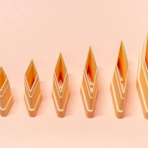 May include: Six geometric, diamond-shaped cookie cutters in varying sizes, arranged from smallest to largest. The cutters are a light peach color with a contrasting cream-colored trim, set against a soft peach background. These are ideal for baking.