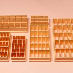 May include: A collection of six beige, rectangular organizers with multiple compartments. The organizers vary in size and shape, with a grid-like interior. The items are arranged on a light pink surface, providing a clean and organized aesthetic.