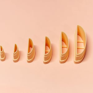May include: A collection of six light orange, semi-circular cookie cutters in ascending sizes. Each cutter has a rounded top, flat base, and a horizontal bar inside. The cutters are displayed against a plain, light peach background.