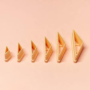 May include: Six light orange, geometric triangle-shaped cookie cutters in a row. The cutters have a hollow centre and vary in size. The design is layered. The smallest cutter is about 2.5 cm tall, the largest is about 7.5 cm.