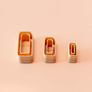 May include: Three beige rectangular cookie cutters of different sizes are displayed on a light pink surface. The cutters have a layered design, with a smaller rectangle inside a larger one. The background is a solid, soft pink.