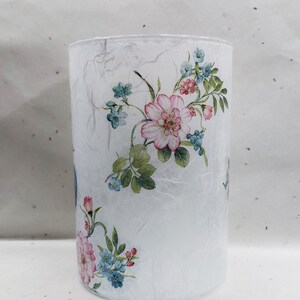 May include: A white glass cylinder vase with a floral design. The vase has a textured surface and features pink and blue flowers with green leaves.