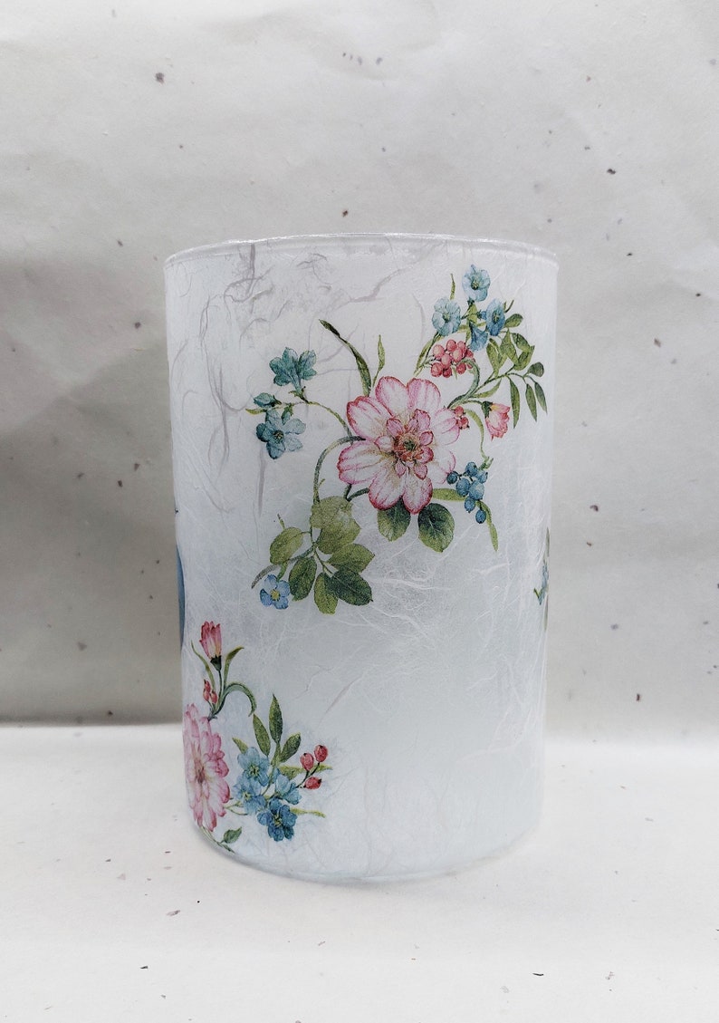 May include: A white glass cylinder vase with a floral design. The vase has a textured surface and features pink and blue flowers with green leaves.