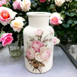 May include: White ceramic vase with a textured surface, decorated with a floral design of pink roses and green leaves.
