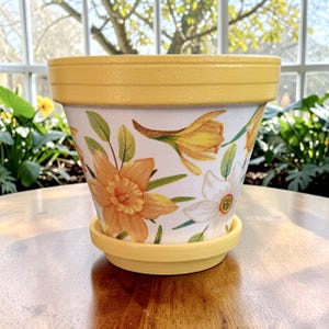 May include: A yellow and white painted flower pot with a matching saucer. The pot is decorated with watercolor-style daffodils in shades of yellow, orange, and white, with green leaves. The pot is sitting on a wooden surface.