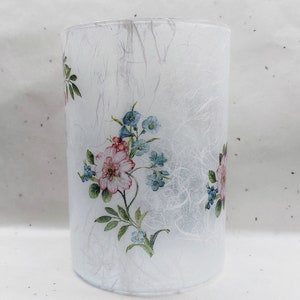 May include: A white glass cylinder vase with a floral design featuring pink and blue flowers. The vase has a textured, white paper-like surface.