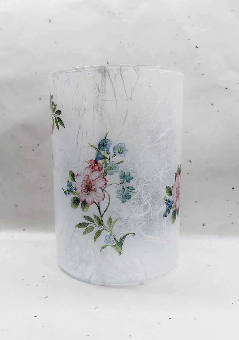 May include: A white glass cylinder vase with a floral design featuring pink and blue flowers. The vase has a textured, white paper-like surface.