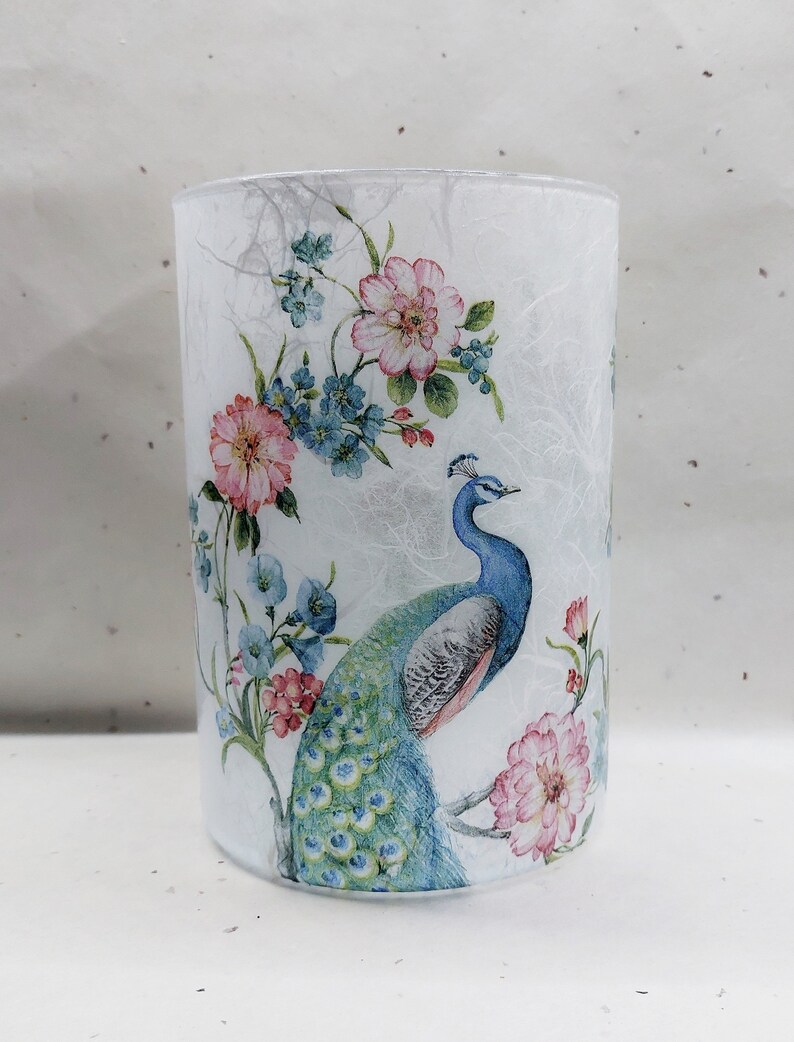 May include: A white glass candle holder with a floral and peacock design. The design features pink, blue, and green colors.