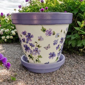 May include: A white terracotta flower pot with a lavender glaze and a matching saucer. The pot is decorated with a hand-painted design of purple flowers and a butterfly.