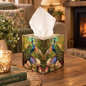 May include: A square tissue box cover featuring a vibrant, symmetrical peacock design with blue and green plumage, surrounded by colourful floral patterns including tulips and hydrangeas, with a white tissue extending from the top opening.